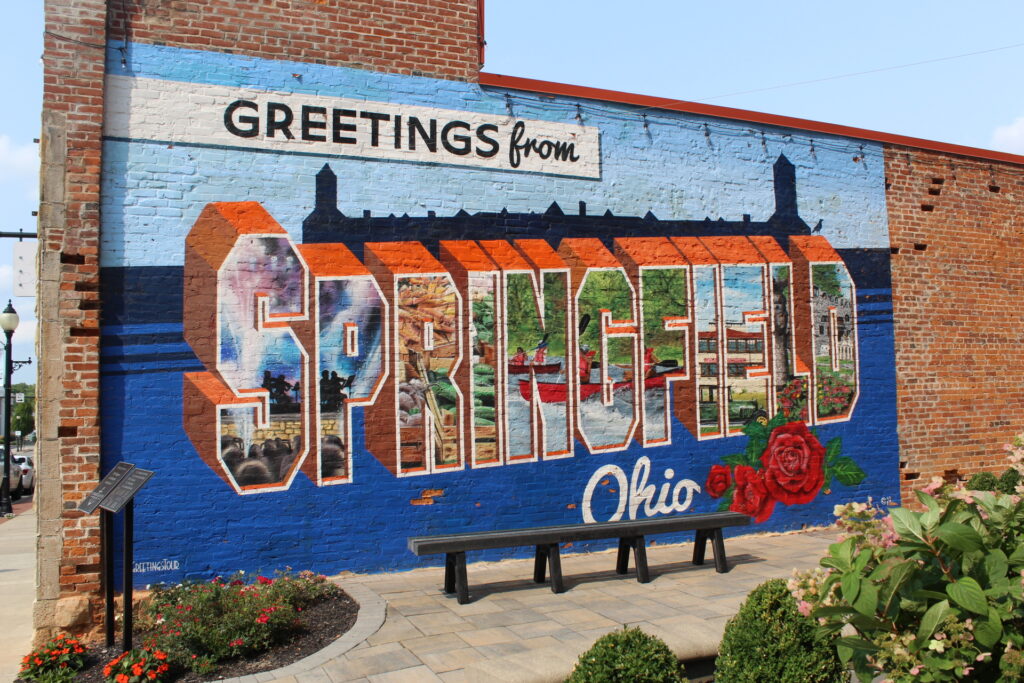 A brick building wall painted with a large mural reading "GREETINGS from SPRINGFIELD Ohio" in postcard style.