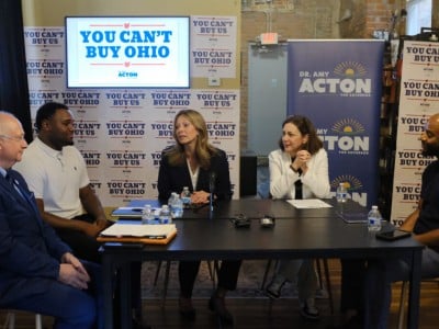 Amy Acton speaks at a panel table surrounded by signs reading "YOU CAN'T BUY OHIO" and "DR. AMY ACTON FOR GOVERNOR."