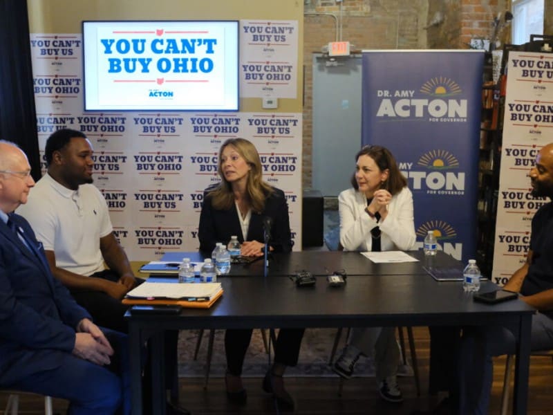 Amy Acton speaks at a panel table surrounded by signs reading "YOU CAN'T BUY OHIO" and "DR. AMY ACTON FOR GOVERNOR."