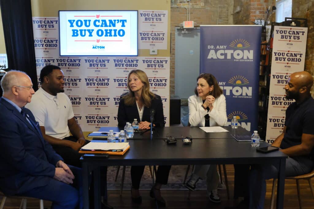 Amy Acton speaks at a panel table surrounded by signs reading "YOU CAN'T BUY OHIO" and "DR. AMY ACTON FOR GOVERNOR."