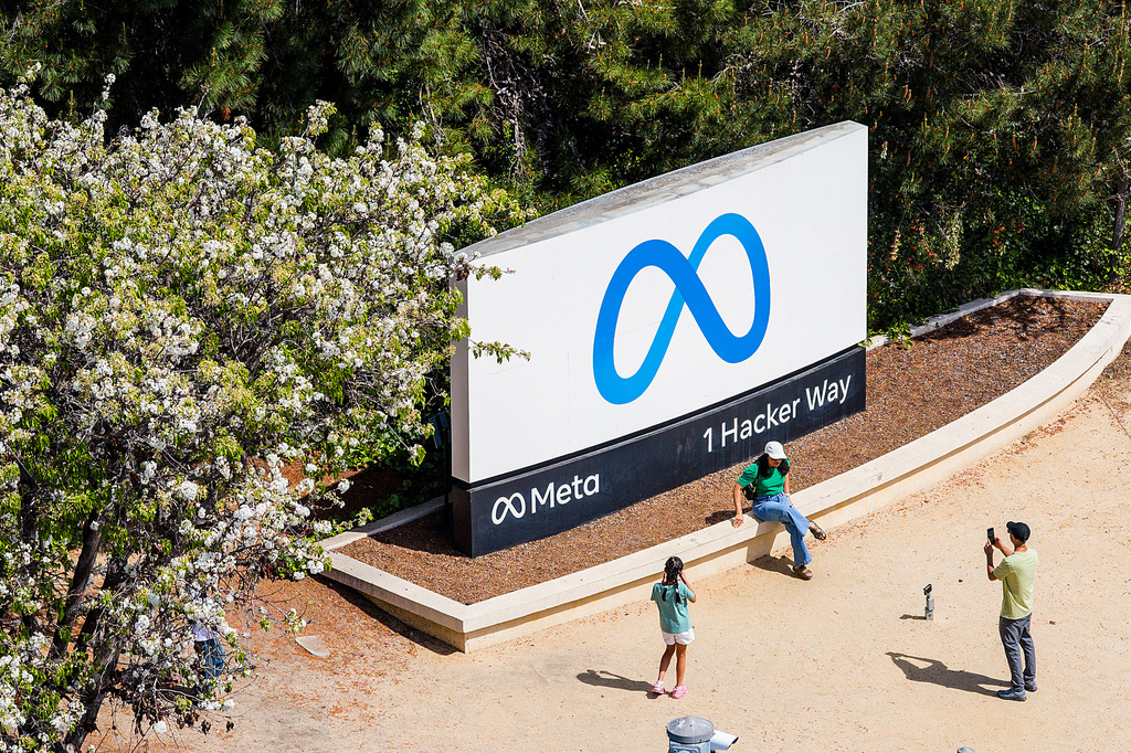 People take photos in front of a large Meta headquarters sign that reads "Meta 1 Hacker Way."