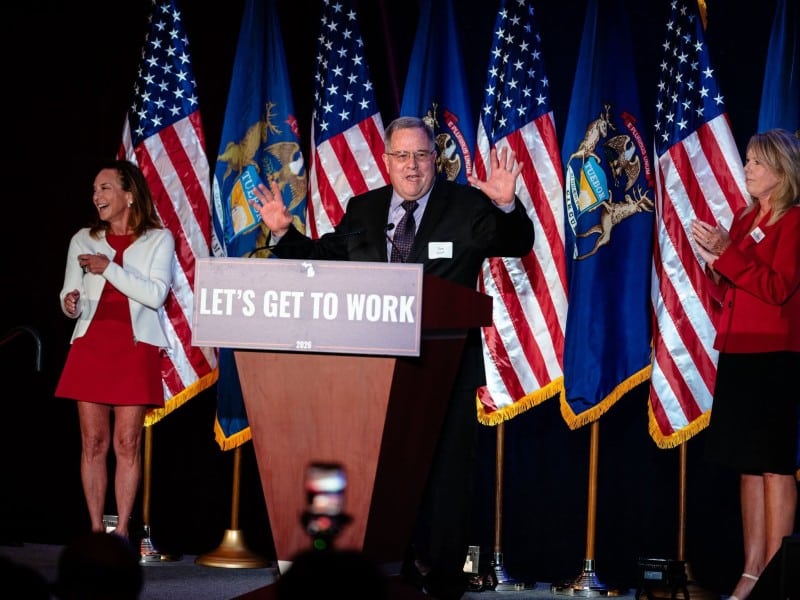 Tom Smith speaks with hands raised at a podium reading "LET'S GET TO WORK" in front of American and Michigan flags.