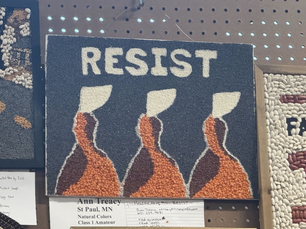 Seed art depicting three figures in red cloaks and white bonnets beneath the word "RESIST."