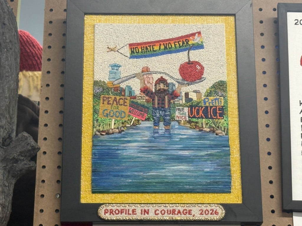 Seed art of Paul Bunyan surrounded by protest signs and a "NO HATE / NO FEAR" banner, labeled "PROFILE IN COURAGE, 2026."