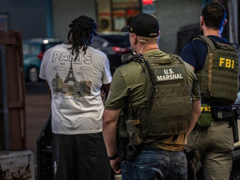 A U.S. Marshal and an FBI agent in tactical gear stand behind a man in a graphic t-shirt.