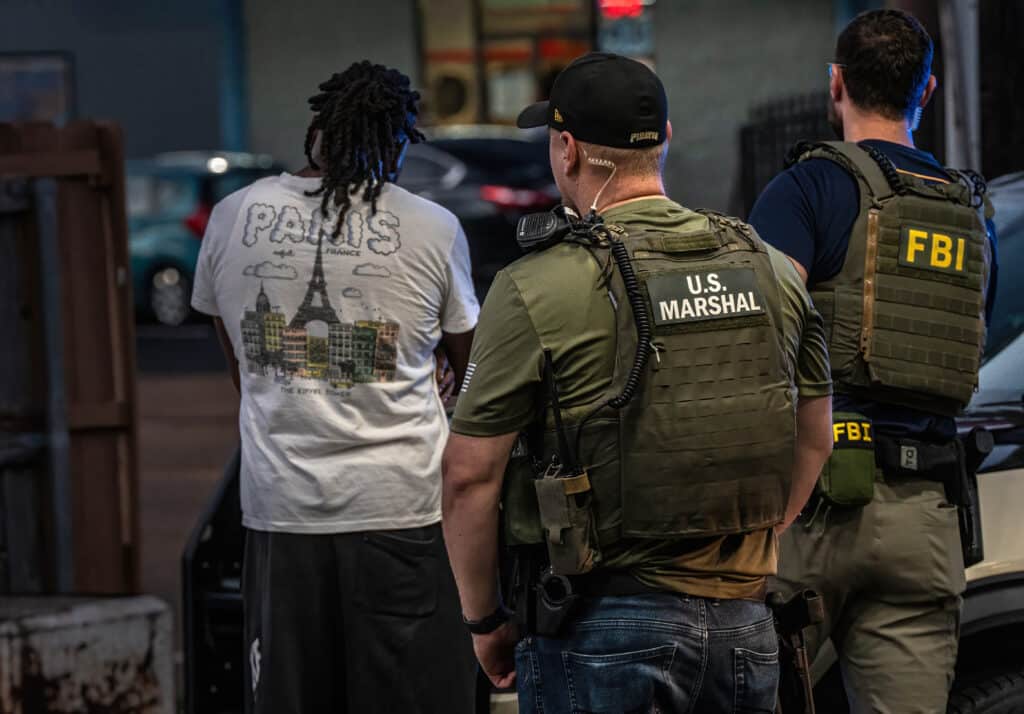 A U.S. Marshal and an FBI agent in tactical gear stand behind a man in a graphic t-shirt.
