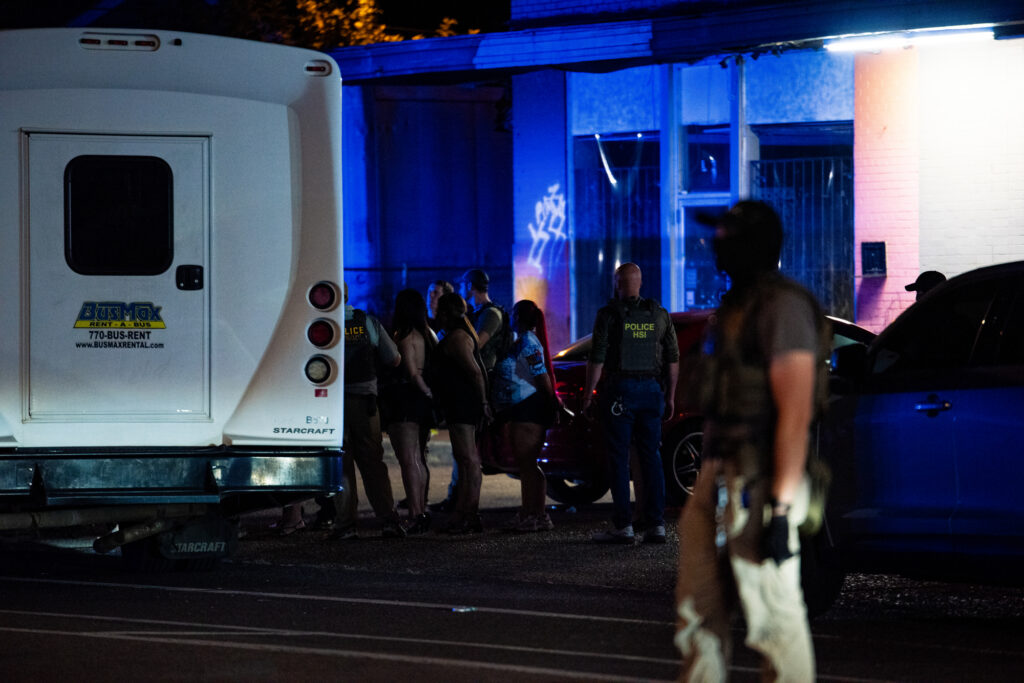 Law enforcement officers stand near a group of people lined up outside a building at night next to a white transport bus.
