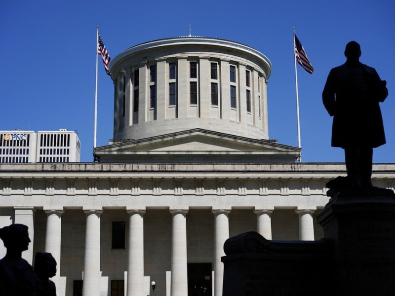 The silhouette of the William McKinley Monument and statues frame the Ohio Statehouse and its rotunda under a clear, blue sky.
