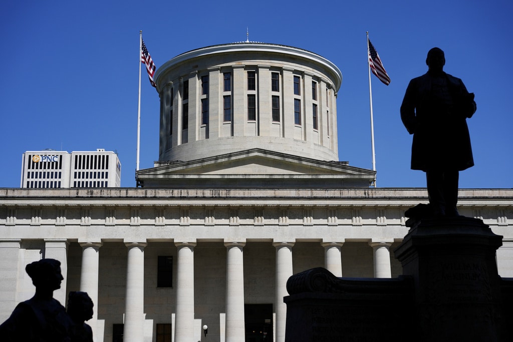 The silhouette of the William McKinley Monument and statues frame the Ohio Statehouse and its rotunda under a clear, blue sky.
