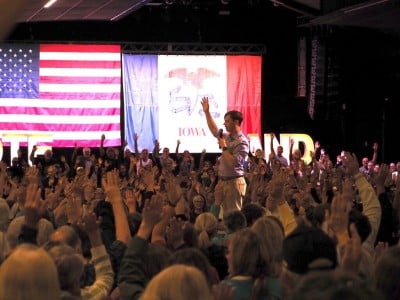 Rob Sand stands on stage with his arm raised as a large crowd of audience members raise their hands in front of American and Iowa flags.