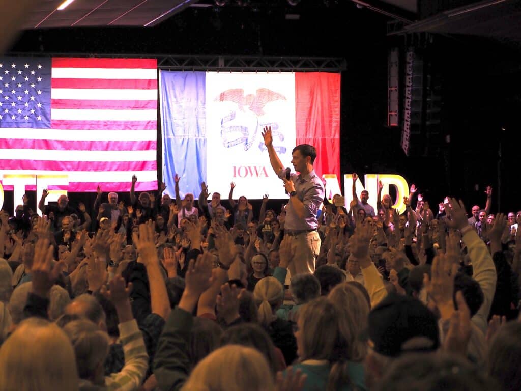 Rob Sand stands on stage with his arm raised as a large crowd of audience members raise their hands in front of American and Iowa flags.