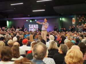 Rob Sand stands onstage with a microphone, gesturing toward a large crowd seated before him at a campaign event.