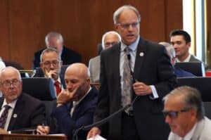 North Dakota House Majority Leader Mike Lefor stands and holds a microphone while speaking in the House chamber.