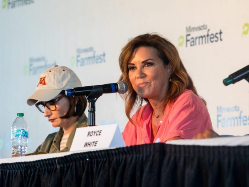 Michele Tafoya speaks into a microphone at a panel table with Farmfest branding behind her.