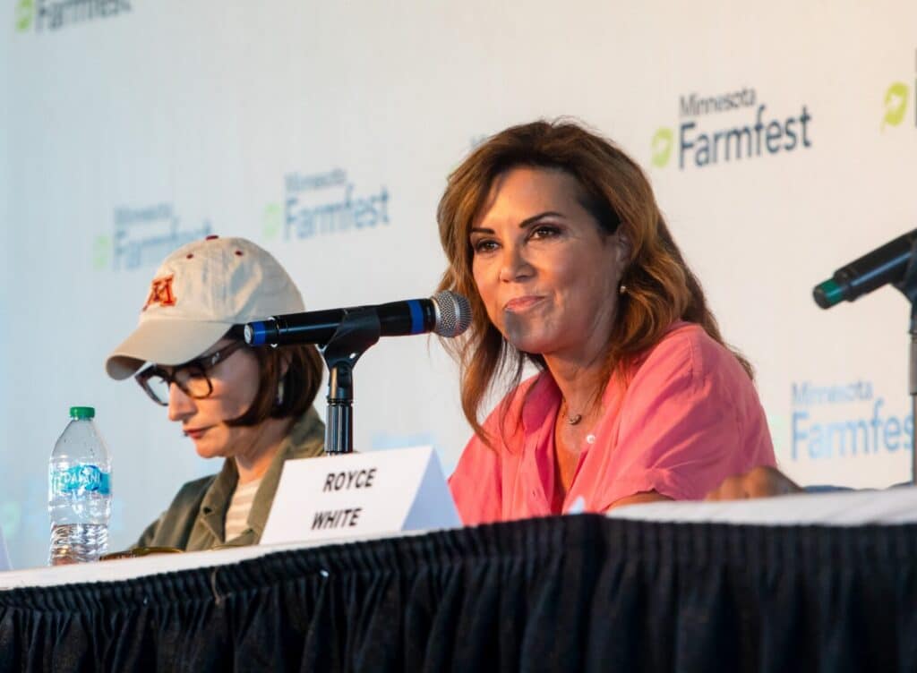 Michele Tafoya speaks into a microphone at a panel table with Farmfest branding behind her.