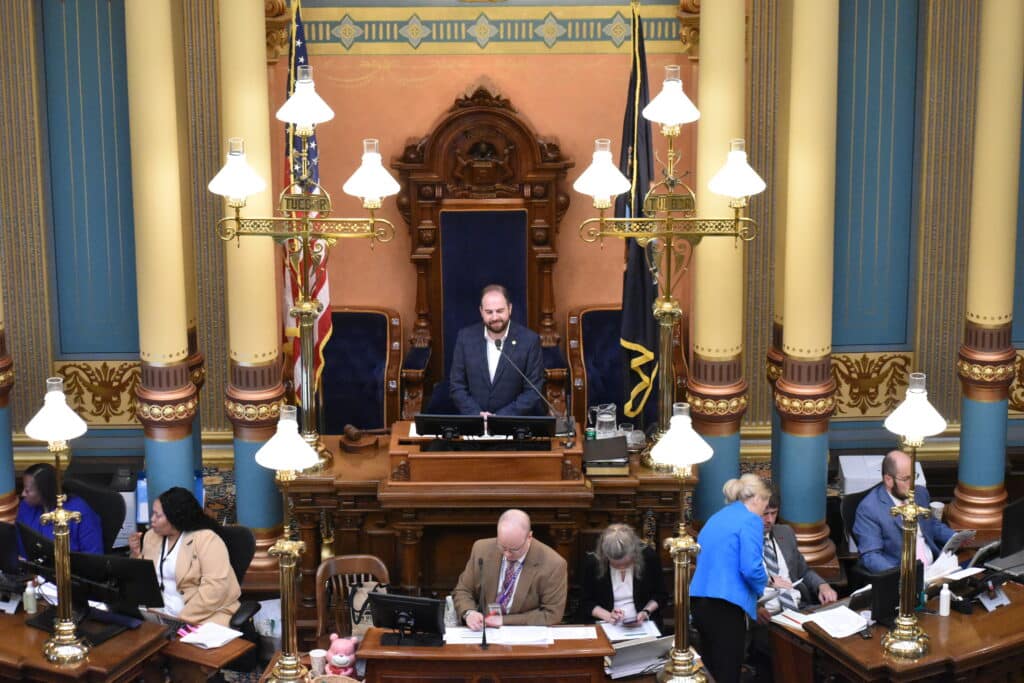 Michigan state Sen. Jeremy Moss stands at the rostrum presiding over a legislative chamber with staff seated below.