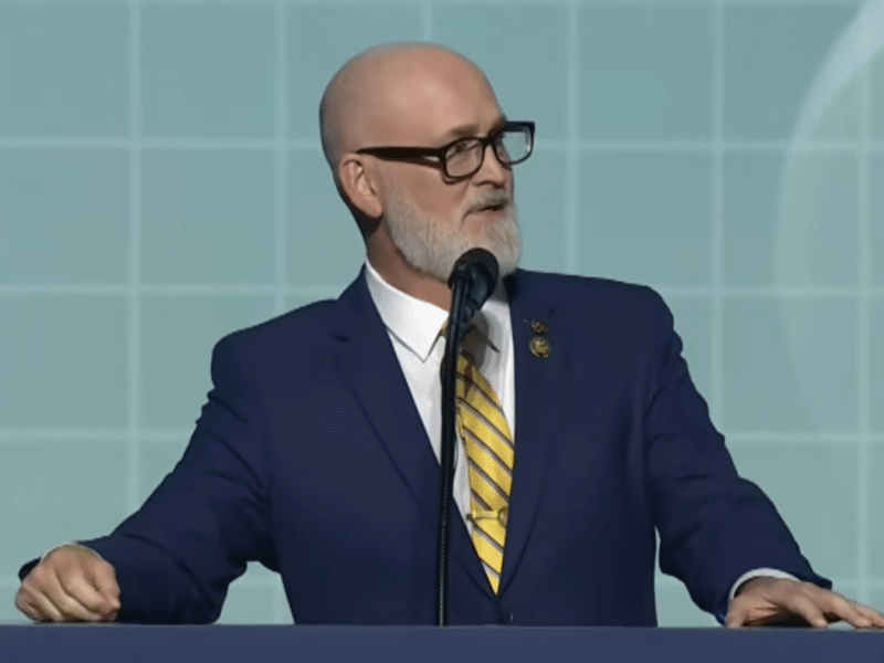 U.S. Rep. Derrick Van Orden, wearing a suit and yellow tie, speaks at a podium against a light blue backdrop.