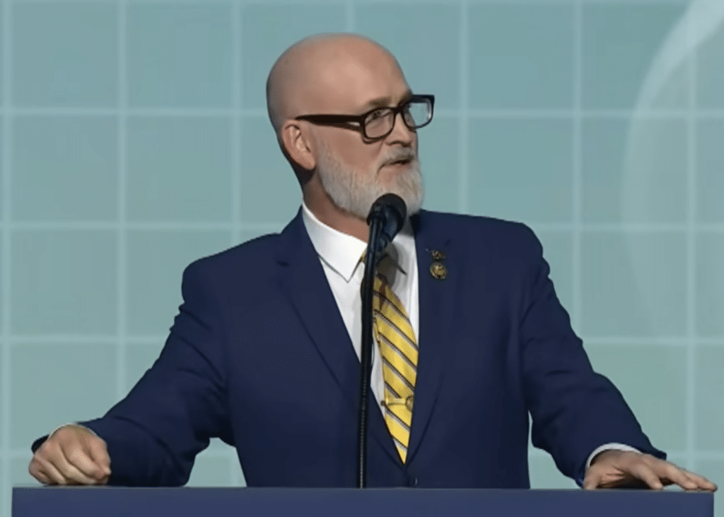 U.S. Rep. Derrick Van Orden, wearing a suit and yellow tie, speaks at a podium against a light blue backdrop.