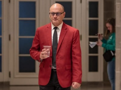 Denny Hoskins, wearing a bright red suit jacket, walks down a hallway holding a plastic cup.