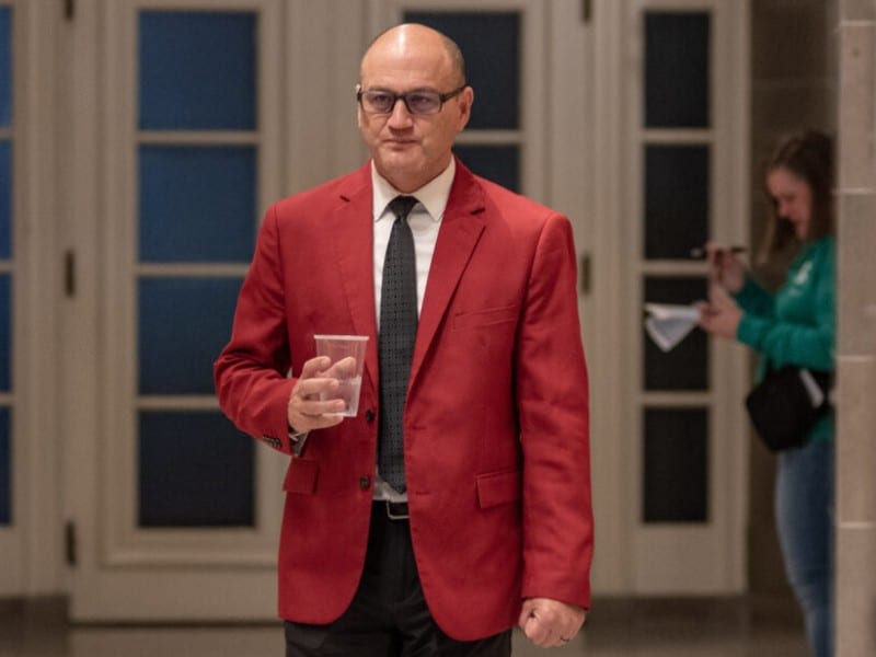 Denny Hoskins, wearing a bright red suit jacket, walks down a hallway holding a plastic cup.