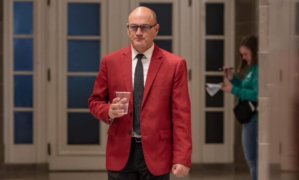 Denny Hoskins, wearing a bright red suit jacket, walks down a hallway holding a plastic cup.