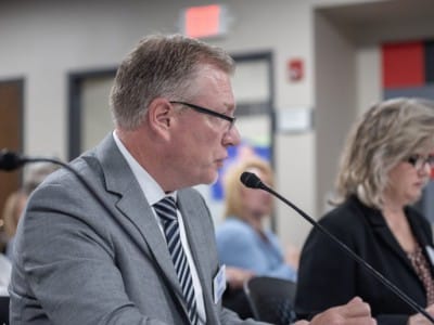Kyle Kruse speaks into a microphone from a side profile while seated at a presentation table.