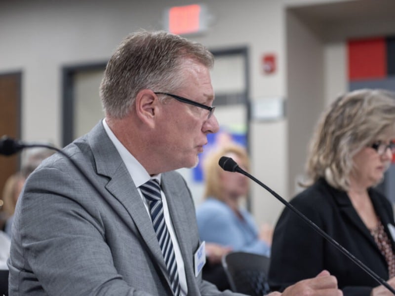 Kyle Kruse speaks into a microphone from a side profile while seated at a presentation table.