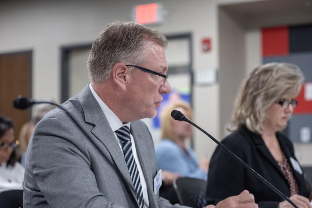 Kyle Kruse speaks into a microphone from a side profile while seated at a presentation table.