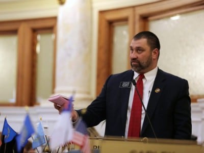 South Dakota state Sen. Tom Pischke speaks into a microphone while holding a small pink booklet on the Senate floor.