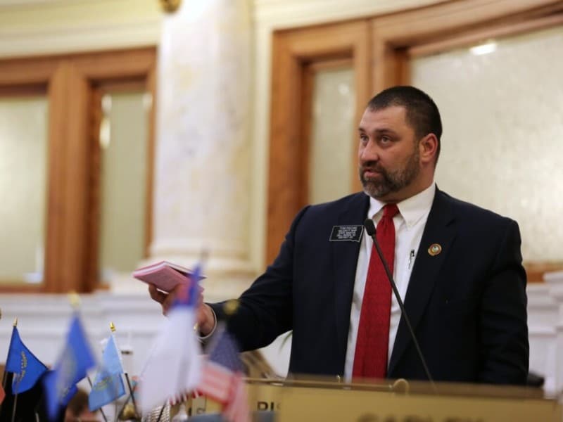 South Dakota state Sen. Tom Pischke speaks into a microphone while holding a small pink booklet on the Senate floor.