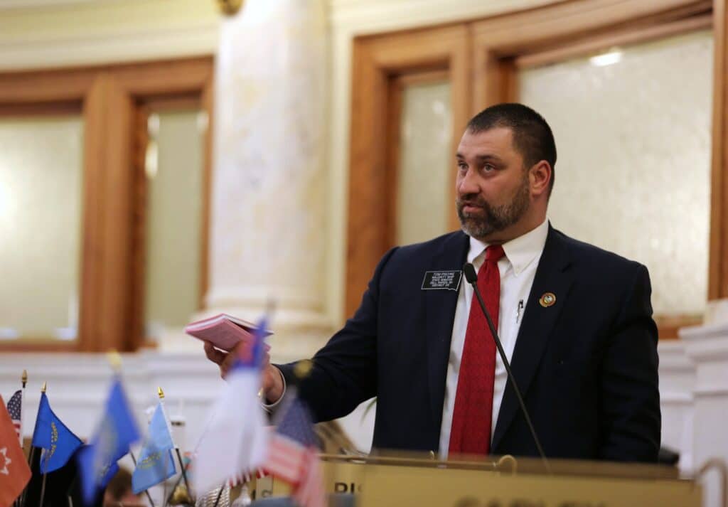 South Dakota state Sen. Tom Pischke speaks into a microphone while holding a small pink booklet on the Senate floor.