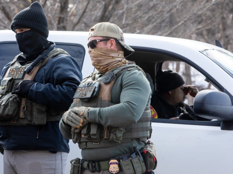 Two masked ICE agents stand outside a pulled-over truck while a man inside pinches the bridge of his nose.