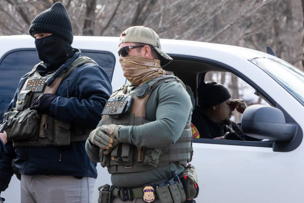 Two masked ICE agents stand outside a pulled-over truck while a man inside pinches the bridge of his nose.