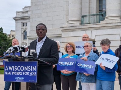 David Crowley speaks at a podium reading "A Wisconsin that works for you," surrounded by supporters with campaign signs.
