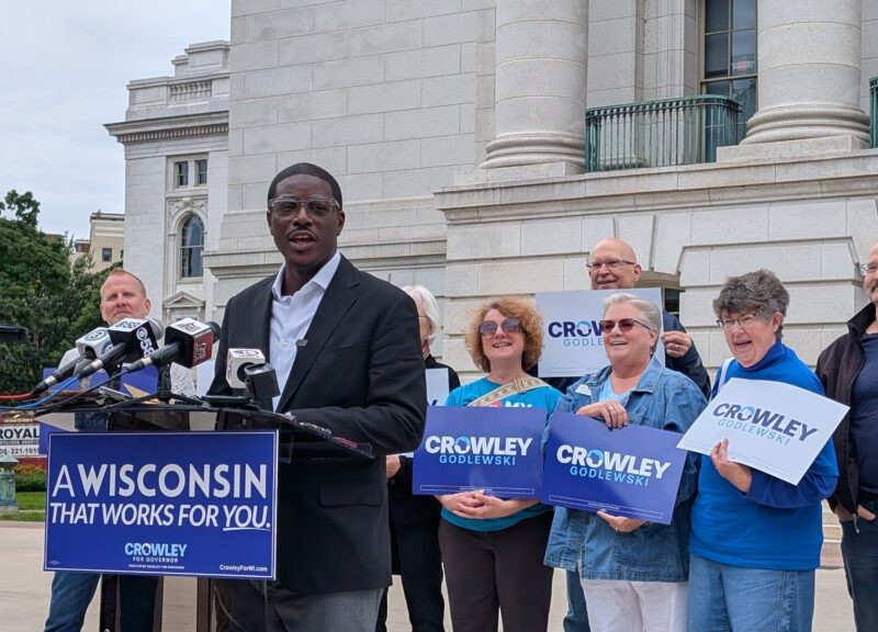 David Crowley speaks at a podium reading "A Wisconsin that works for you," surrounded by supporters with campaign signs.