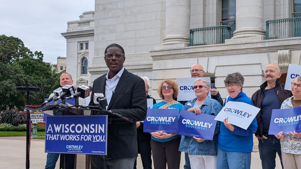 David Crowley speaks at a podium reading "A Wisconsin that works for you," surrounded by supporters with campaign signs.