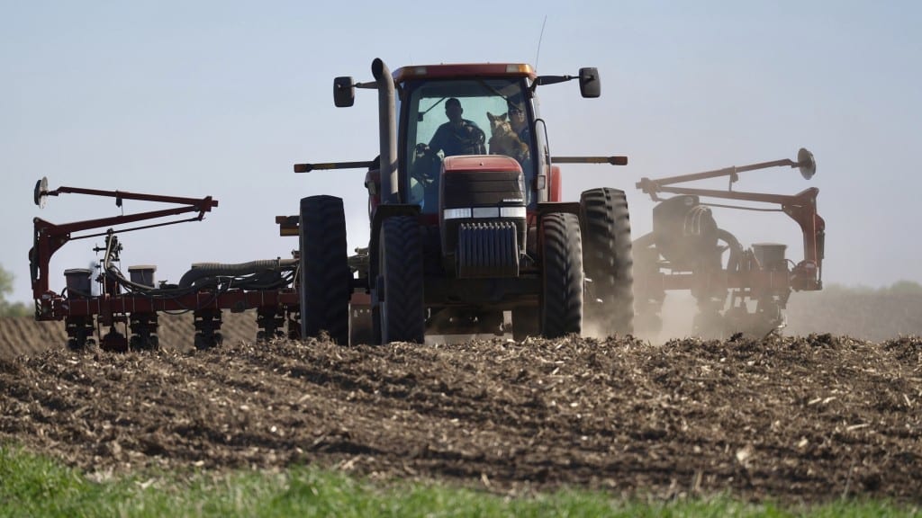A farmer and a dog sit in the cab of a tractor pulling planting equipment across a dirt field.