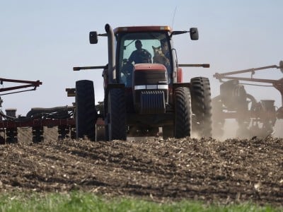 A farmer and a dog sit in the cab of a tractor pulling planting equipment across a dirt field.