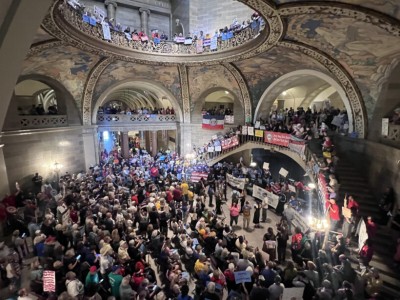 A high-angle view of a crowded, multi-tier Missouri Capitol rotunda filled with protesters holding signs and banners.
