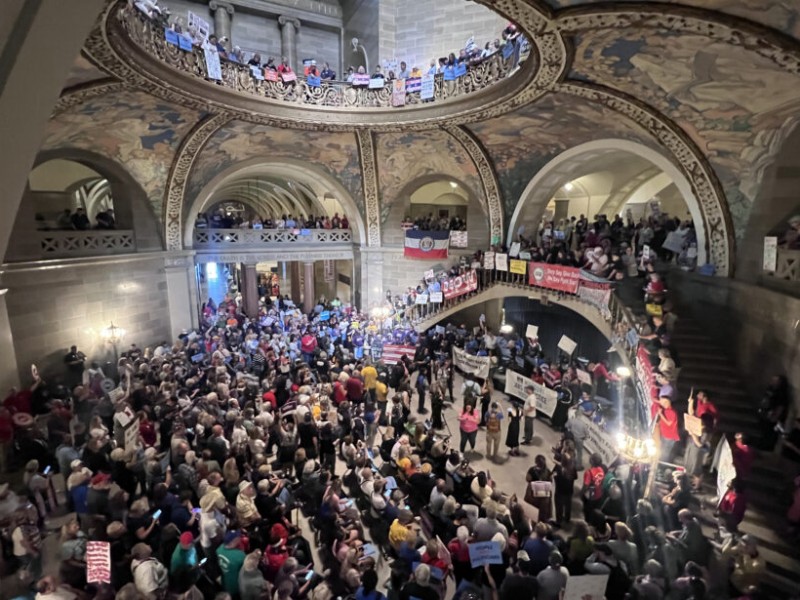 A high-angle view of a crowded, multi-tier Missouri Capitol rotunda filled with protesters holding signs and banners.