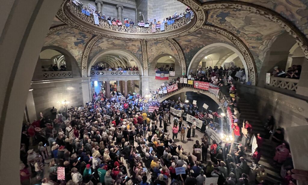 A high-angle view of a crowded, multi-tier Missouri Capitol rotunda filled with protesters holding signs and banners.