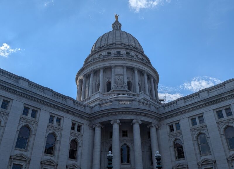 The white stone exterior dome and facade of the Wisconsin State Capitol building against a blue sky.