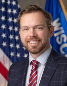 Adam Neylon smiles in an official headshot against the U.S. and Wisconsin state flags.