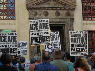 Protesters hold signs in front of a building entrance reading "Justice for Pierre!" and "End the war on our Haitian neighbors now!"