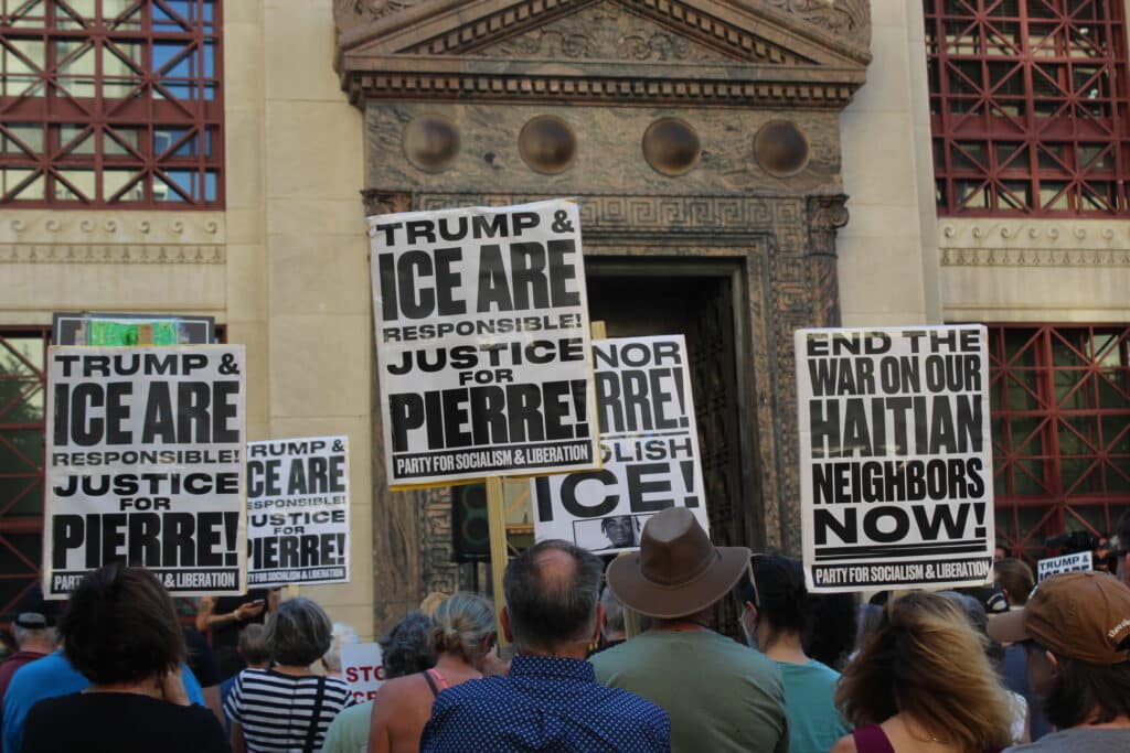 Protesters hold signs in front of a building entrance reading "Justice for Pierre!" and "End the war on our Haitian neighbors now!"