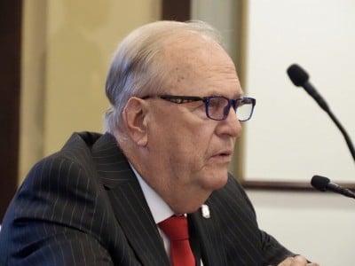 Danny Williams speaks into a microphone during a committee meeting, wearing glasses and a suit.