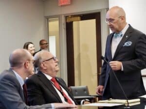 Mike Osburn, wearing a suit and bow tie, stands and speaks to Danny Williams at a committee table.
