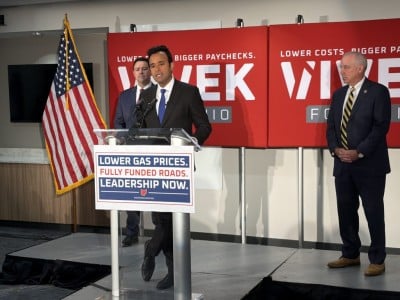 Vivek Ramaswamy speaks at a podium reading "Lower gas prices. Fully funded roads. Leadership now."