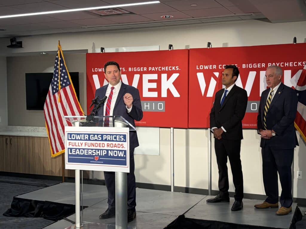 Rob McColley speaks at a podium while Vivek Ramaswamy and Matt Huffman stand beside him on stage.