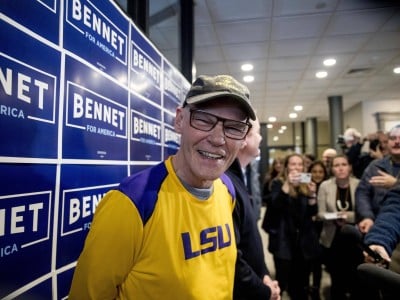 James Carville smiles while standing in front of a wall covered in blue Bennet for America campaign signs.